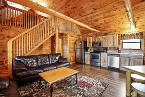 A wooden cabin interior with a leather sofa, wooden staircase, kitchen area, and a dining table. - The Aspen Cabin | Hocking Hills, Charming, Hot Tub (Rockbridge)