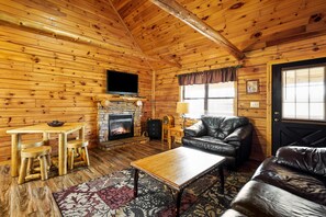 A log cabin interior with a stone fireplace, leather furniture, and a television mounted on the wall. - The Aspen Cabin | Hocking Hills, Charming, Hot Tub (Rockbridge)