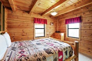 A wooden cabin room with a bed, two windows with red curtains, a dresser, and a ceiling light.