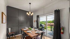 Een eetgedeelte met een houten tafel gedekt voor zes personen, omringd door zwarte stoelen. Een kroonluchter hangt aan het plafond, en een brandblusser zit aan de muur bevestigd.