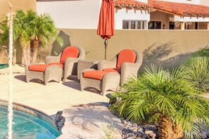 A poolside area with wicker chairs and an umbrella, surrounded by palm trees and a pool.