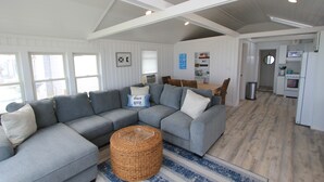 Un salon spacieux avec un grand canapé d’angle gris, un pouf rond en osier et un tapis  à rayures bleues et blanches.