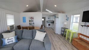 Un vaste espace de vie comprenant un canapé gris, un banc en bois et un téléviseur fixé au mur. On aperçoit une cuisine à l’arrière-plan, avec des meubles blancs et un four à micro-ondes.