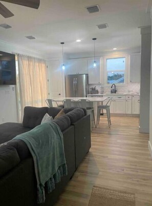 A modern living room with a dark sofa, a TV, and a dining area with a table and chairs. - PCB FUN-heated pool, hot tub, golf cart, putt putt (Panama City Beach)