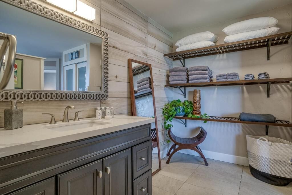 A bathroom with a double sink vanity, a large mirror, shelves with towels, and a plant.