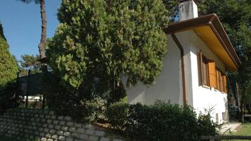 Un pequeño edificio blanco con techo marrón y contraventanas de madera, rodeado de follaje y árboles.