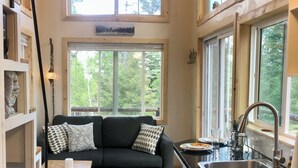 A cozy living room with a dark green sofa, patterned cushions, a small wooden table, and a colorful rug. A large window offers a view of trees.