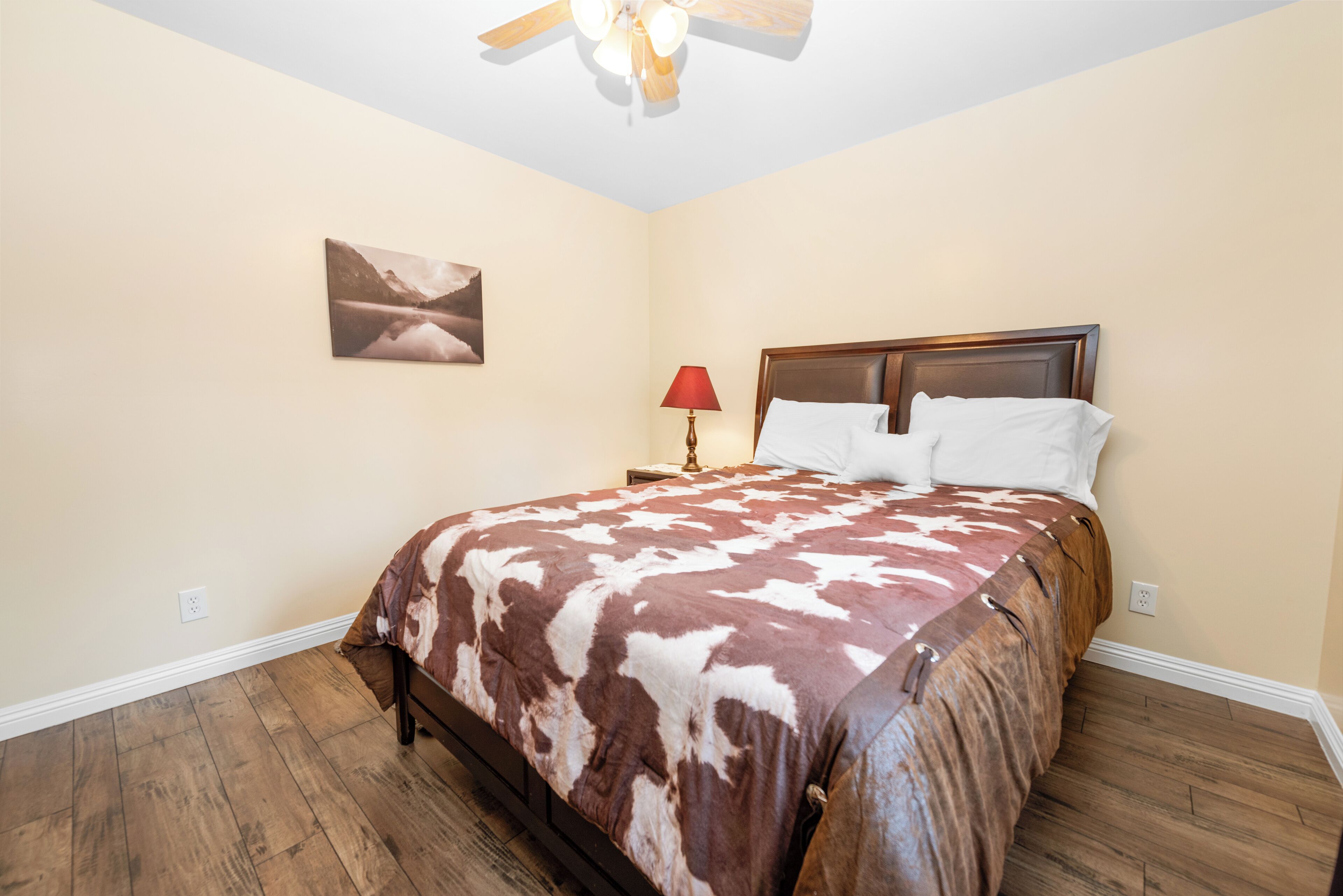 A bedroom with a bed, a ceiling fan, a nightstand with a lamp, and a wall-mounted painting.