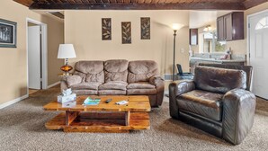 Ein Wohnzimmer mit einer braunen Ledersofa, einem Ledersessel und einem Couchtisch aus Holz. Ein Lampenschirm, ein gerahmtes Bild an der Wand und eine Küchenzeile im Hintergrund sind ebenfalls zu sehen.