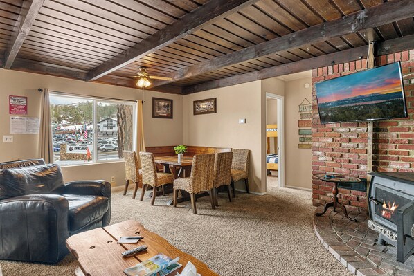 A cozy living room with a fireplace, a dining area with wicker chairs, a leather sofa, and a TV mounted on a brick wall.