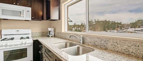 A kitchen with dark wooden cabinets, a white microwave, a stove, a sink, and a window with a view of trees and a snowy landscape.