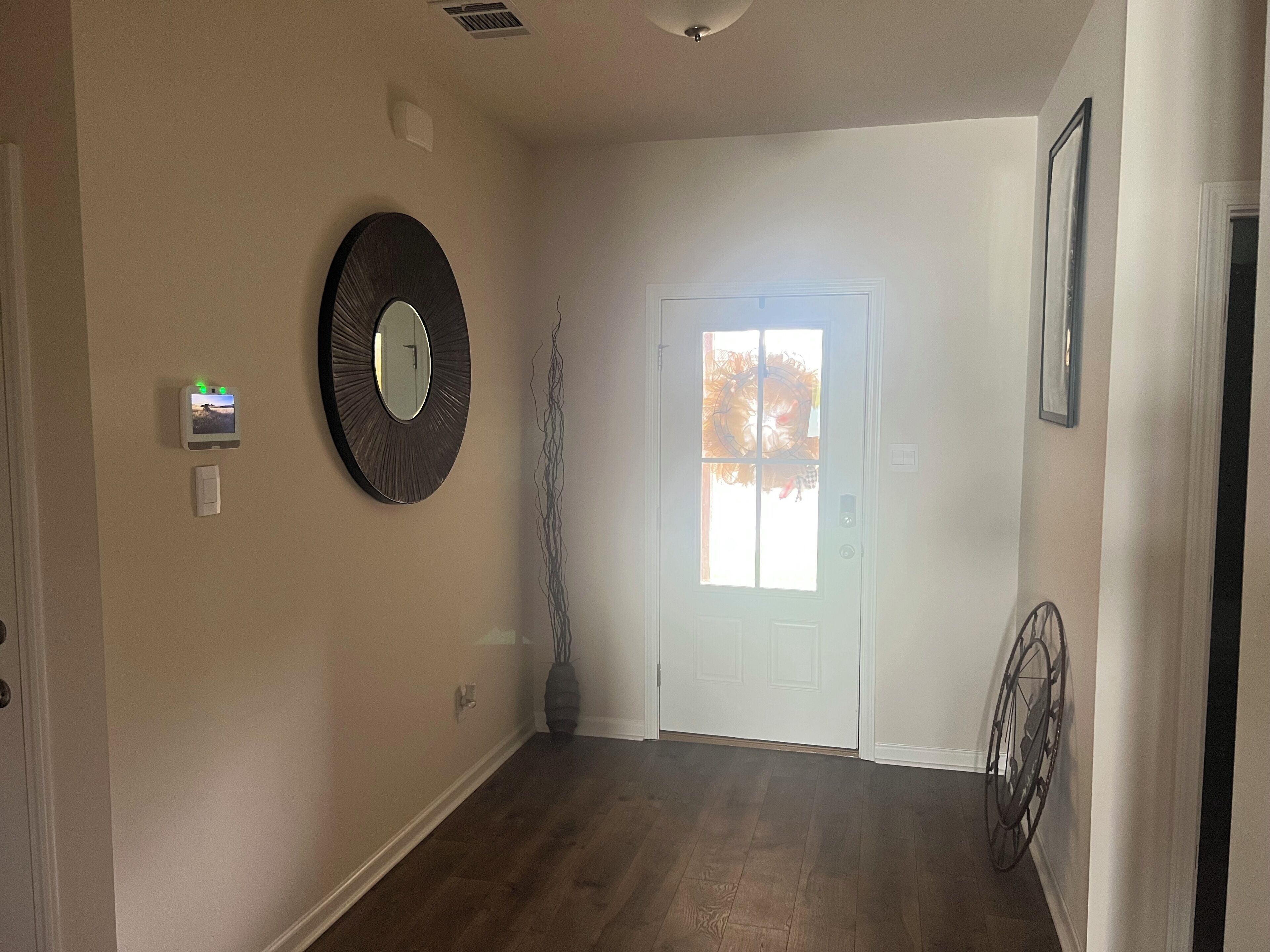 A hallway with a wooden floor, a door with a window, a wall-mounted thermostat, and a decorative mirror.