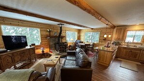A cozy cabin interior with a stone fireplace, wooden furniture, and large windows offering views of the forest. - Cozy two bedroom cabin with hearty Firewood Stove/Game Room (Springville)