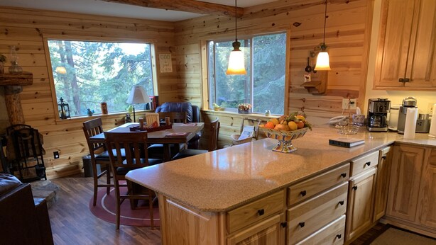 A cabin interior with a wooden dining area, a kitchen with granite countertops, and large windows offering a view of the forest. - Cozy two bedroom cabin with hearty Firewood Stove/Game Room (Springville)