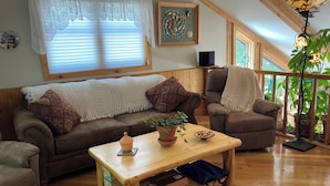 Uma sala de estar aconchegante com um sofá de couro marrom, uma mesa de centro de madeira e uma poltrona marrom. Há uma janela com cortinas brancas, um quadro na parede e um vaso com planta sobre a mesa de centro.