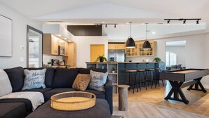 A modern living room with a sofa, a coffee table, and a pool table. There is a kitchen area with a bar and stools in the background.