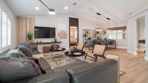 A spacious living room with a large sofa, a dining area with a table and chairs, and a kitchen area with a TV and plants.