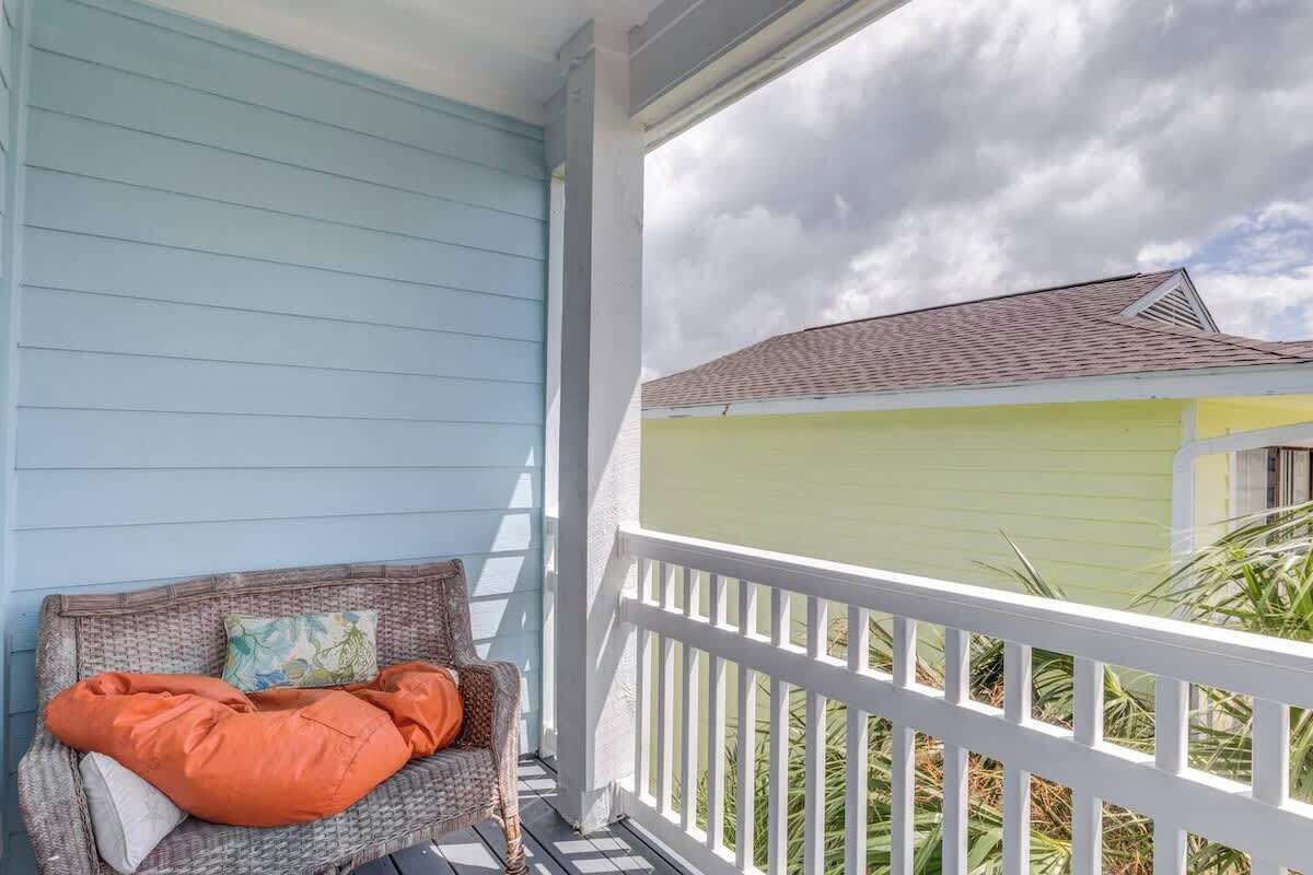 A porch with a wicker chair and cushions, a white railing, and a view of a house with a green roof.