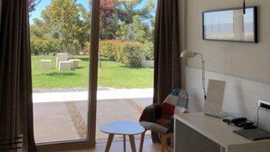 Ein modernes Hotelzimmer mit einer großen Schiebeglasdoor, die zu einem Garten führt, einem kleinen runden Tisch, einer Couch und einem Schreibtisch mit einem Laptop.
