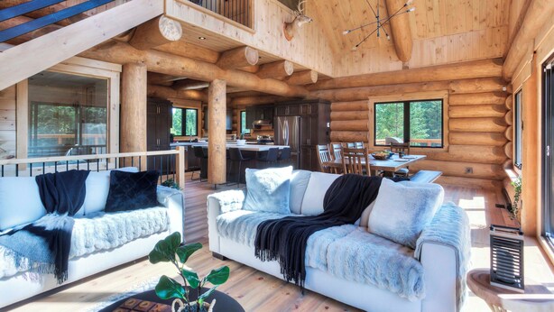 Een ruim houten blokhutinterieur met een eethoek, keuken en een groot raam met uitzicht op het bos.