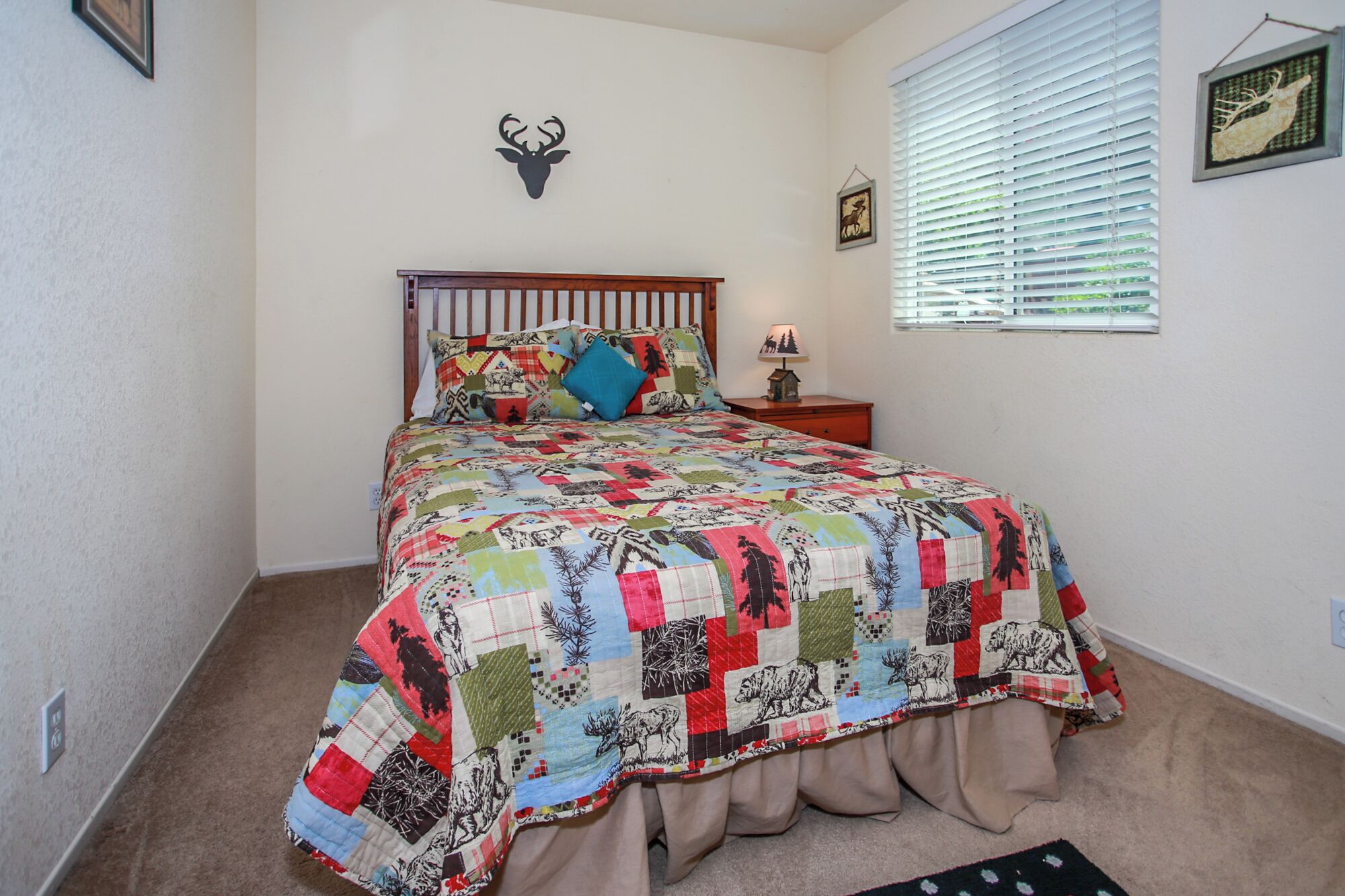 Un dormitorio con cama de madera, colcha colorida, decoración de ciervo en la pared, mesita de noche con lámpara y ventana con persianas.