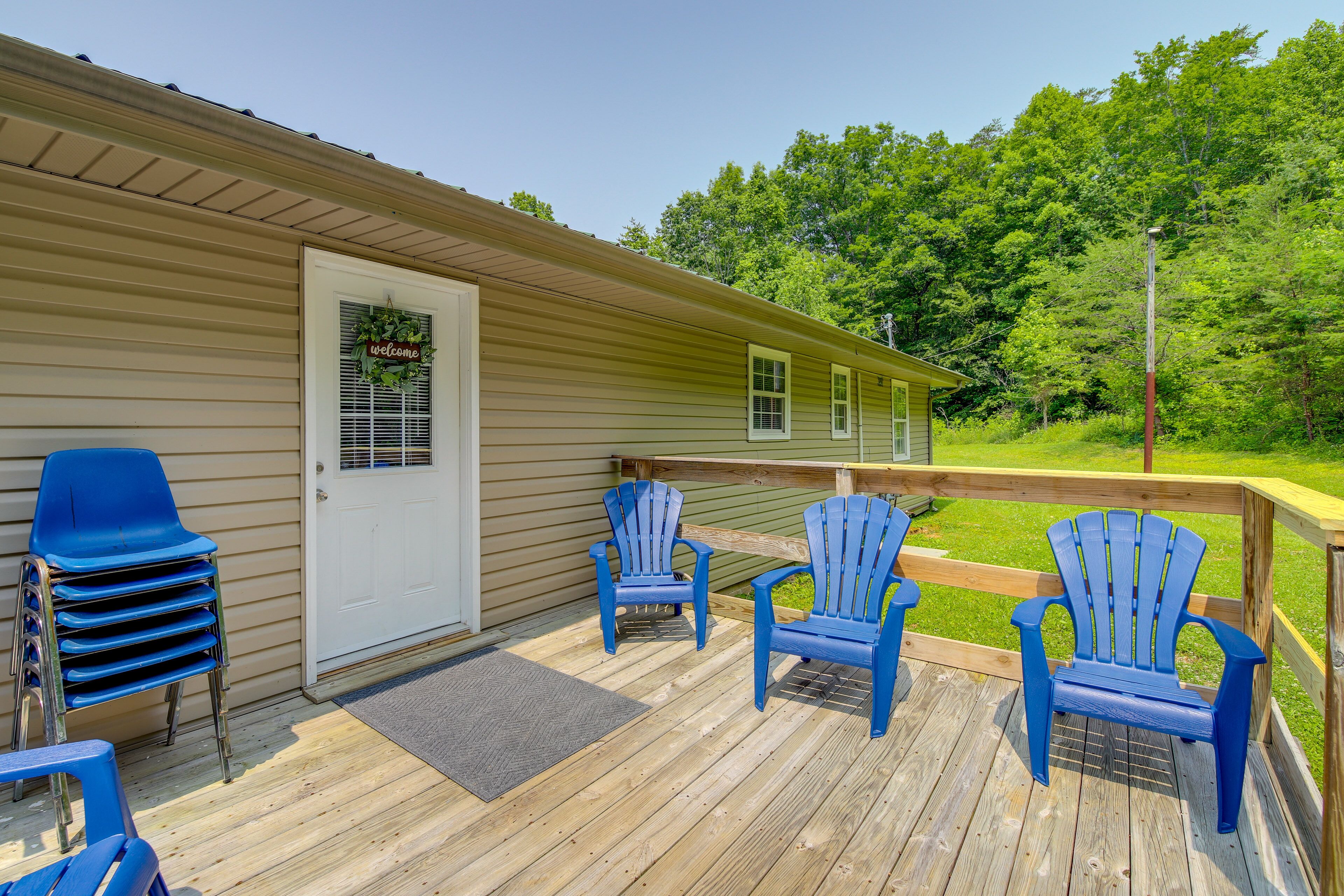 A wooden deck with blue Adirondack chairs, a door with a 'Welcome' sign, and a view of a green landscape.