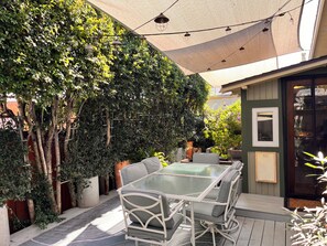 En uteplats med ett glasbord och stolar, omgiven av frodig grönska och en pergola.