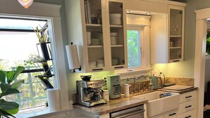 A modern kitchen with white cabinets, a built-in oven, and a coffee machine. There is a dining table with chairs and a window with plants. - Urban Greenhouse (San Diego)