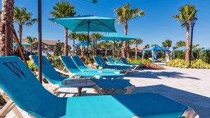 Zona junto a la piscina con sillas reclinables azules y sombrillas, rodeada de palmeras y un cielo despejado.