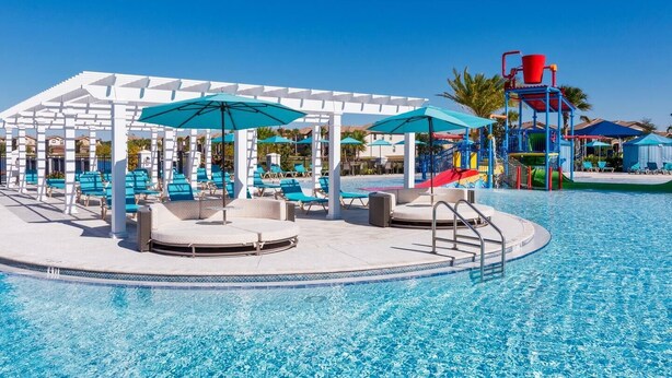 Un área de piscina con sillones de descanso y sombrillas, un tobogán acuático y un solárium.