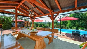 A covered outdoor dining area with wooden tables and benches, a pool in the background, and red umbrellas. - Nice home in Vinogradi Ludbreski with sauna (Vinogradi Ludbreski)