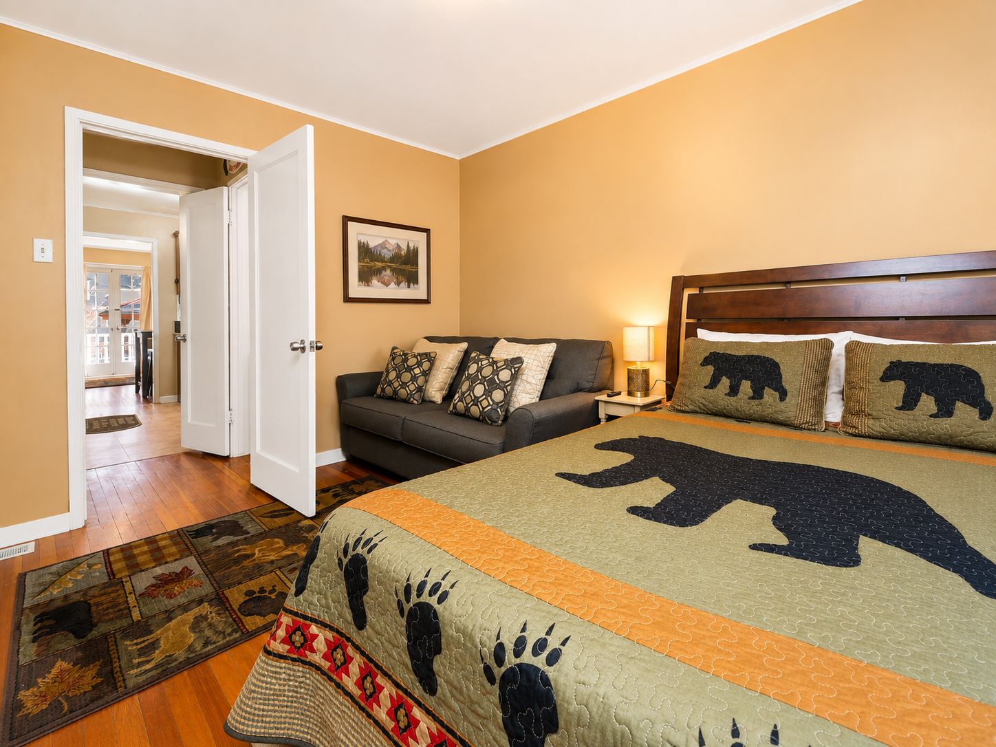 Quarto com cama, sofá, mesa de cabeceira, abajur, tapete e porta que dá para outro cômodo.