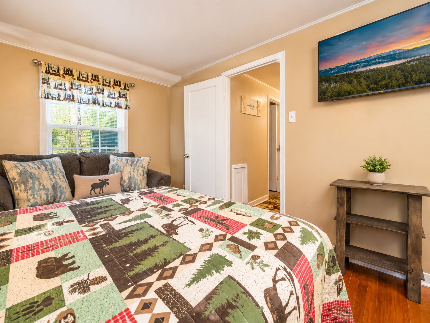 A bedroom with a large bed, a window with curtains, a door, a painting on the wall, and a small plant on a shelf.