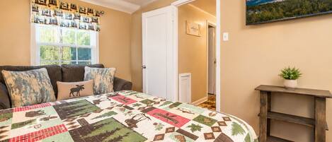 A bedroom with a large bed, a window with curtains, a door, a painting on the wall, and a small plant on a shelf.