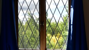 Une fenêtre ornée d’une grille à motifs losangés, avec des rideaux, et donnant sur des arbres.