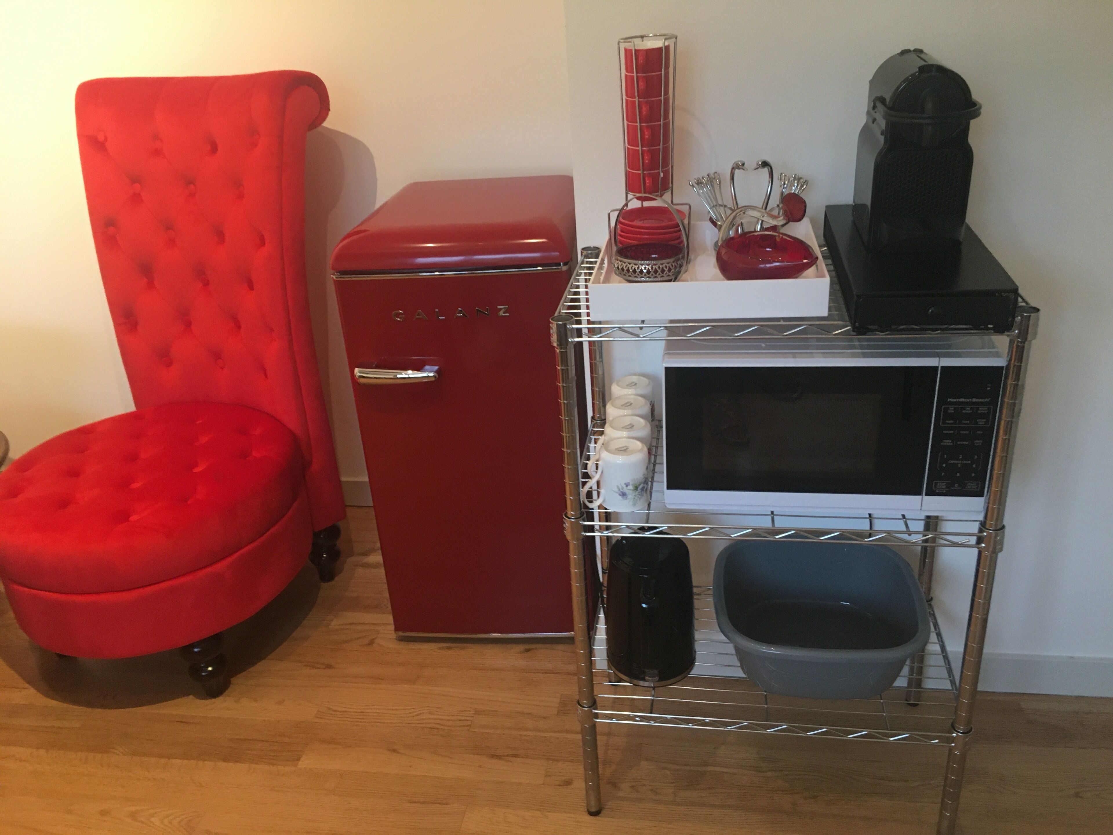 A compact kitchenette with a red refrigerator, a microwave, and a small shelf holding kitchenware.
