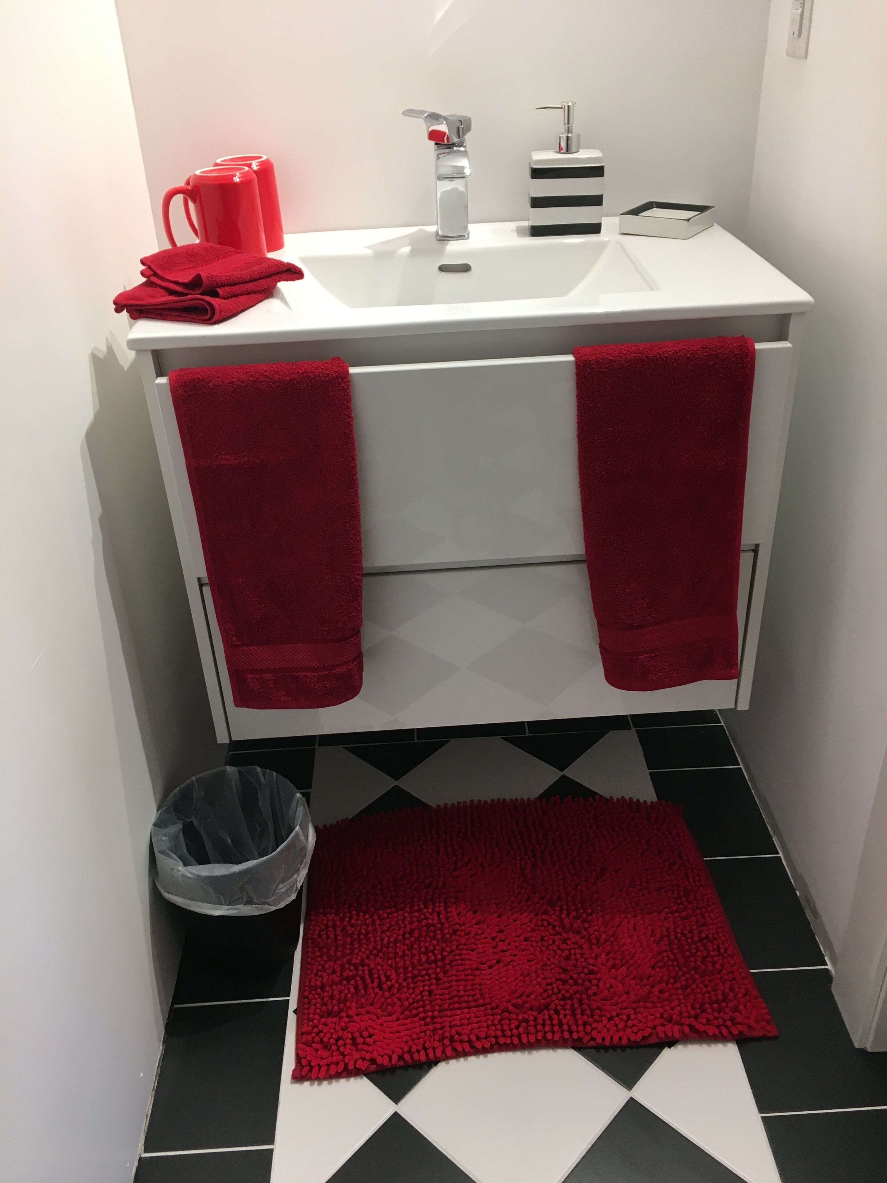 Une salle de bain avec un lavabo blanc, des serviettes rouges, un tapis rouge et une poubelle.