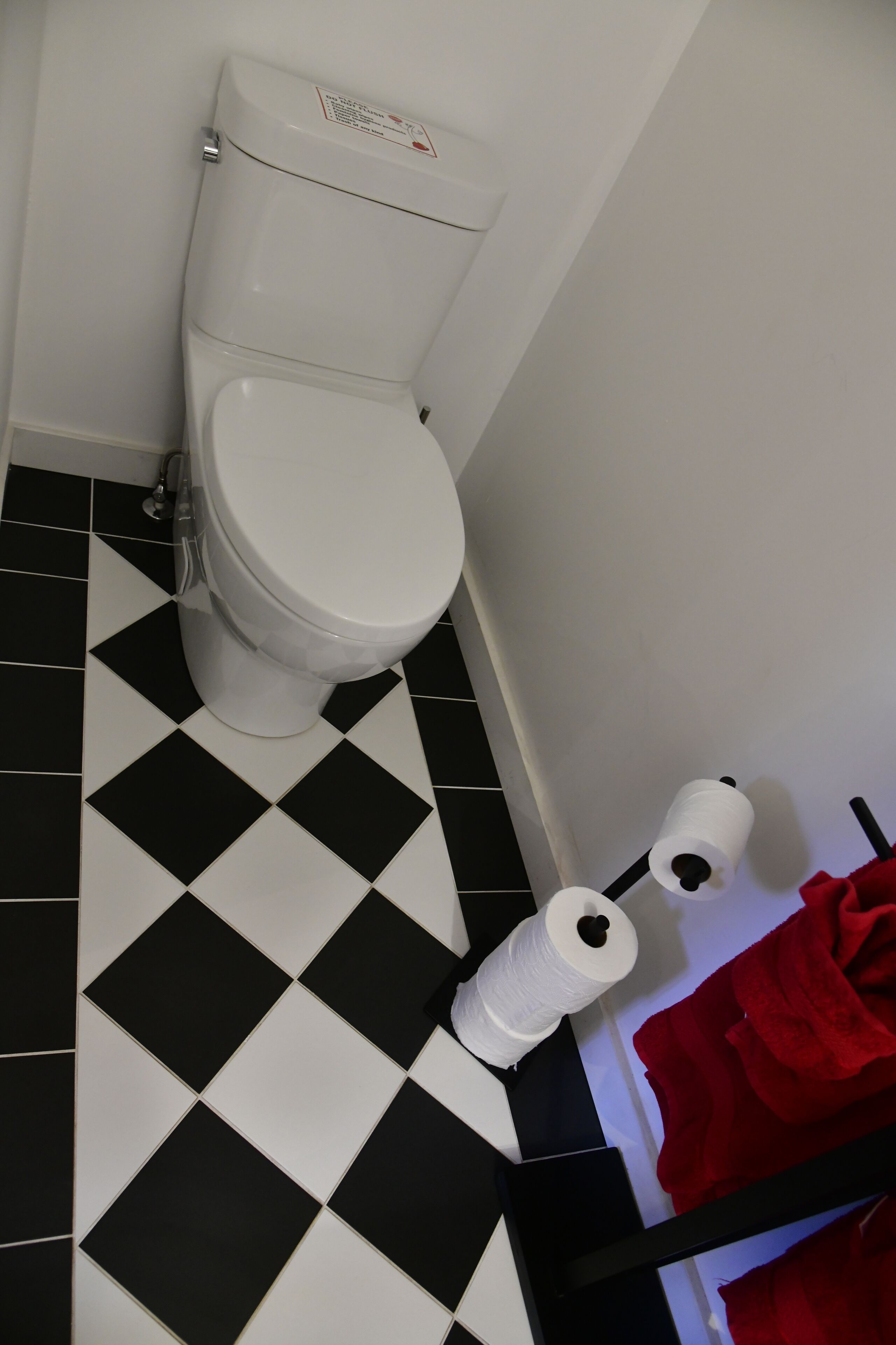 Een toilet met een zwart-wit schaakkleed vloerpatroon, een witte toiletrolhouder, en een rode handdoek op een rek.