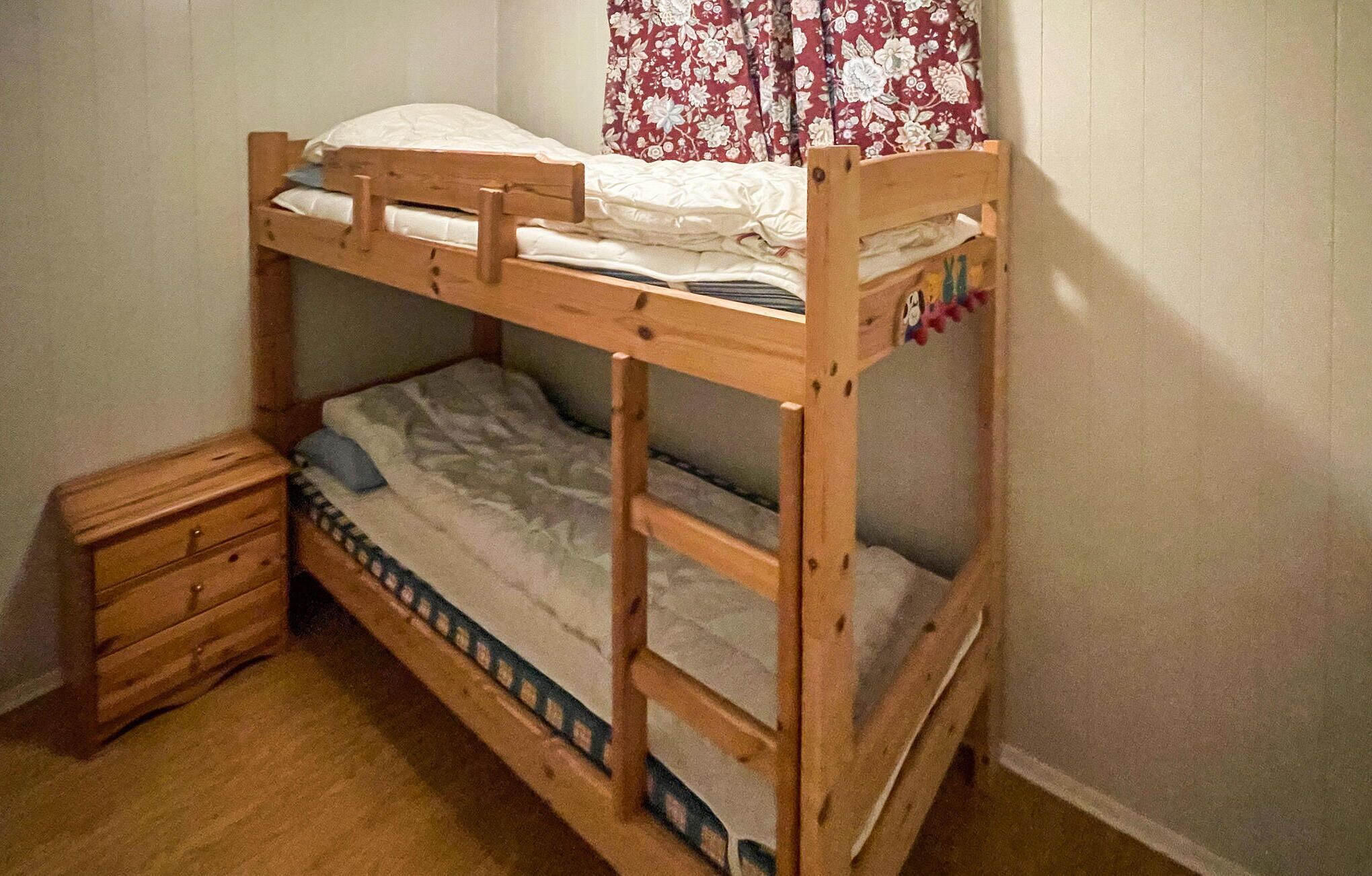 Un lit superposé avec une structure en bois, un lit inférieur recouvert d’une couverture à motifs et un lit supérieur avec un drap blanc. Un petit meuble en bois avec des tiroirs se trouve à gauche du lit.