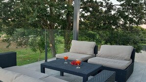 Un área de estar al aire libre con mobiliario de mimbre y una mesa de vidrio, rodeada de vegetación.