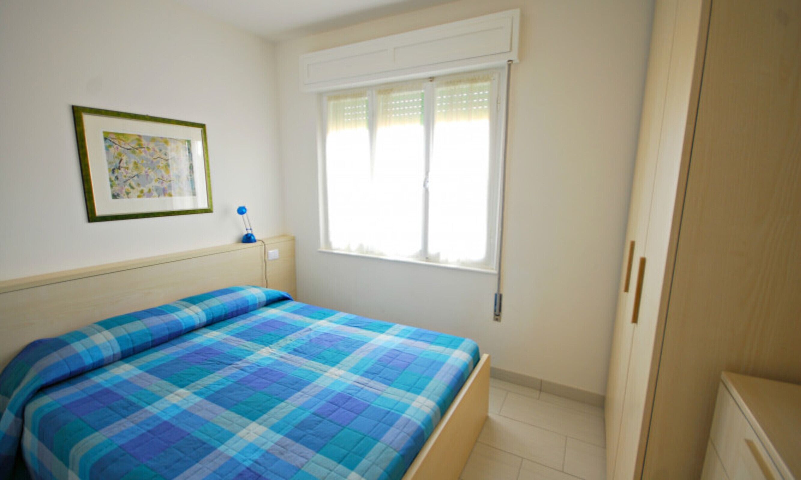 Un dormitorio con cama, una ventana con persianas, una mesita de noche con lámpara y un cuadro enmarcado en la pared.
