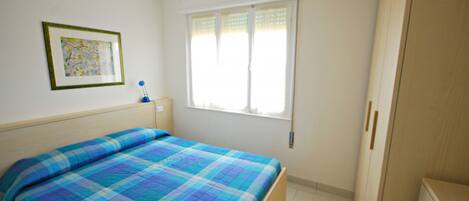 Un dormitorio con cama, una ventana con persianas, una mesita de noche con lámpara y un cuadro enmarcado en la pared.