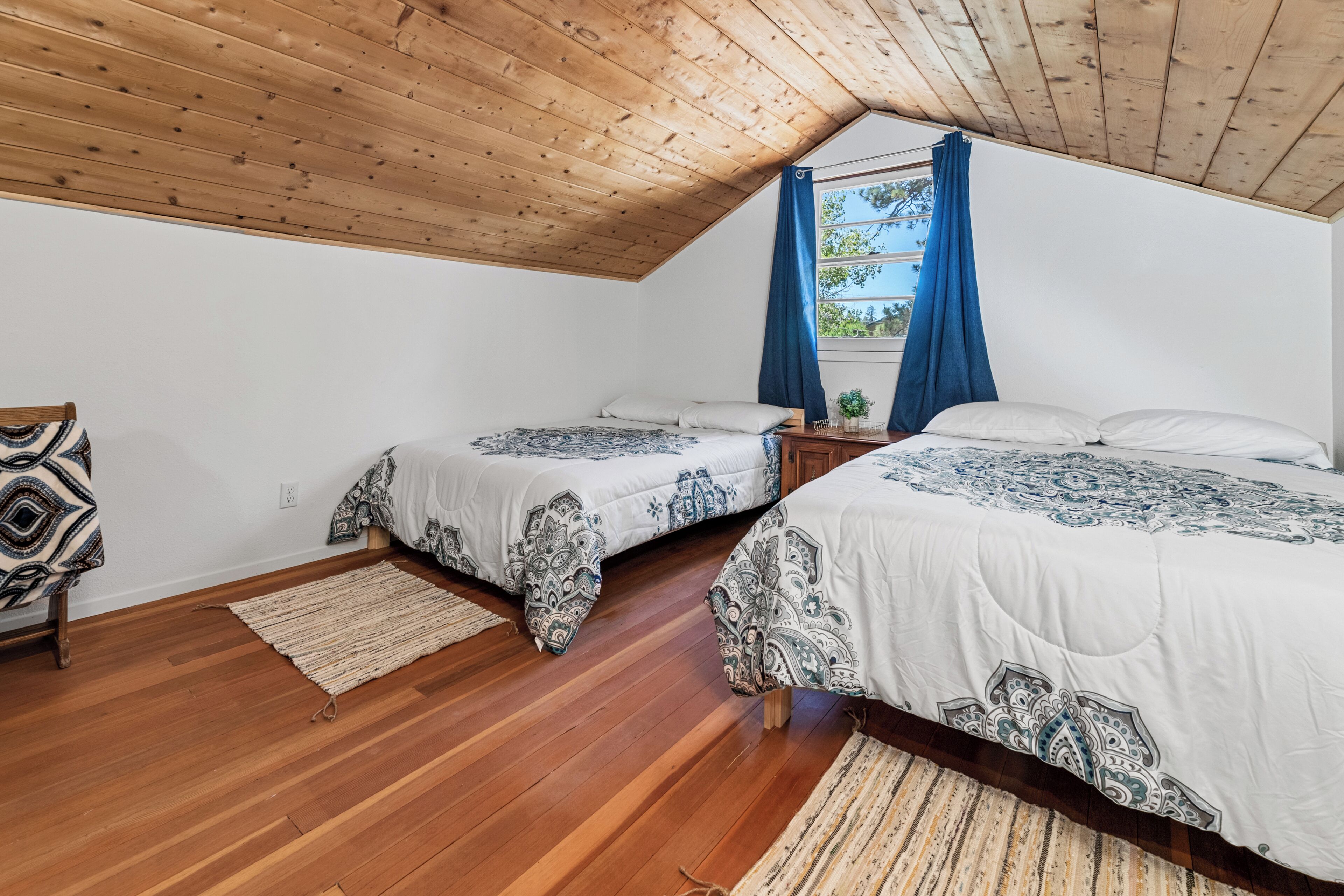 A bedroom with two beds, wooden flooring, a sloped ceiling, and a window with blue curtains.