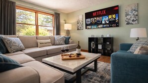 Una sala de estar con un sofá, una mesa de centro con copas de vino y una botella, una pared con televisión y carteles de Netflix, y una estantería.