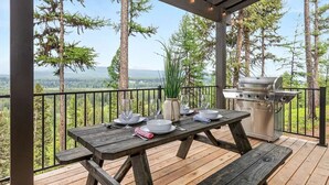 Ein Essbereich im Freien mit einem Holztisch, gedeckt für vier Personen, einem Grill und Blick auf eine bewaldete Landschaft.