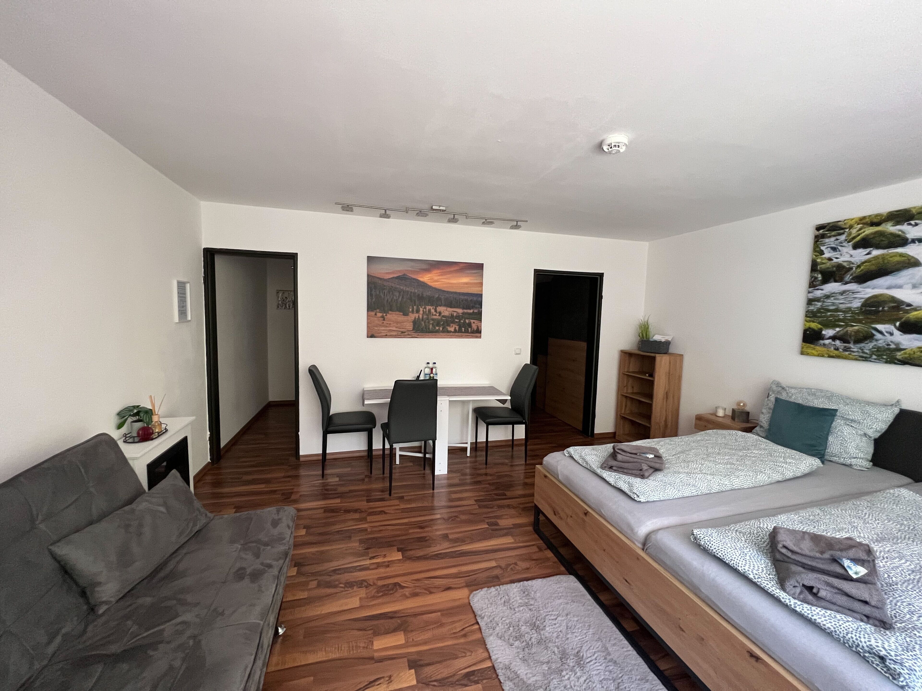 Ein kompakter Raum mit Bett, Schlafsofa, Schreibtisch und Stuhl. An der Wand hängt ein Bild, und eine Deckenleuchte erhellt den Raum.