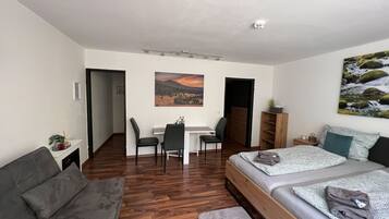Ein kompakter Raum mit Bett, Schlafsofa, Schreibtisch und Stuhl. An der Wand hängt ein Bild, und eine Deckenleuchte erhellt den Raum.