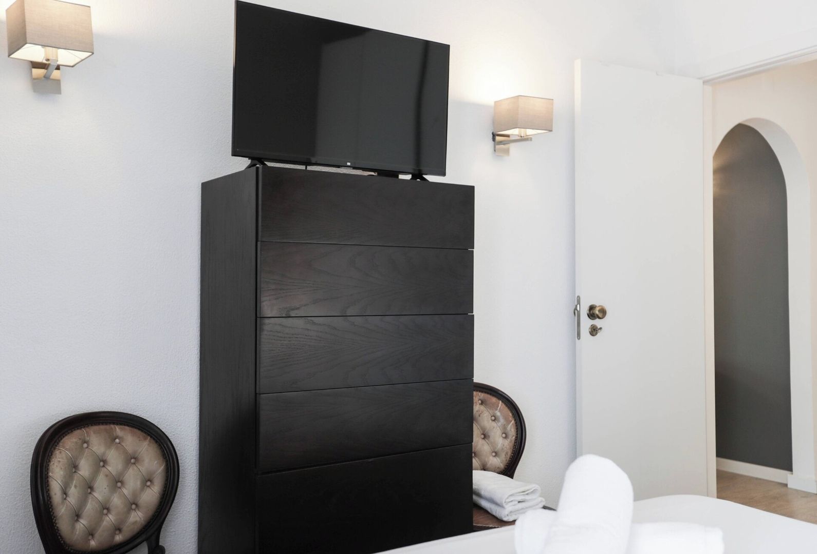 A hotel room with a large wardrobe, a bed with white linens, two upholstered chairs, a television mounted on the wardrobe, and a door with an arched frame.