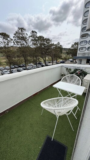 Une terrasse sur le toit, recouverte d’un gazon synthétique, avec deux chaises en métal blanc, une petite table et, en arrière-plan, un bâtiment blanc doté de fenêtres rondes.
