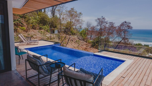 A modern outdoor pool area with a view of the ocean, wooden deck, and lounge chairs.
